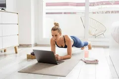 Kobieta ćwiczy plank w domu, patrząc na laptop podczas treningu online.